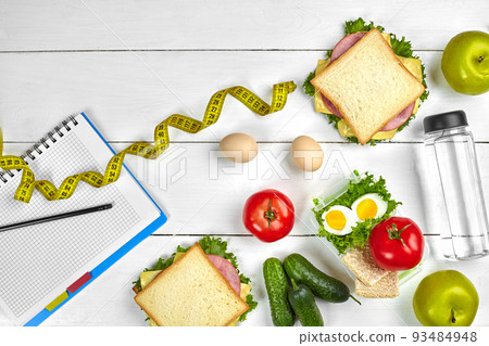 Top view planning notebook with copy space and healthy breakfast. Sandwich with ham and cheese, eggs, cucumber, tomato and bottle of water on white wooden table. Top view planning notebook with copy space and healthy breakfast. Sandwich with ham and cheese, eggs, cucumber, tomato and bottle of water on white wooden table. 93484948