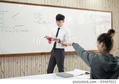 An Asian male teacher in a classroom near the whiteboard is writing and teaching a lesson with a special lecture in a class column. Math teacher explains arithmetic sums to university students An Asian male teacher in a classroom near the whiteboard is writing and teaching a lesson with a special lecture in a class column. Math teacher explains arithmetic sums to university students 93487021