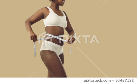 Slender woman who loses weight measures her waist with measuring tape standing on beige background. Slender woman who loses weight measures her waist with measuring tape standing on beige background. 93487158