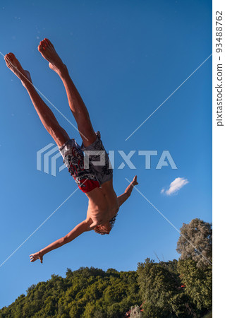Young teen boy jumping flying and diving in the river. Clear blue sky and trees in distance as a natural background. 93488762
