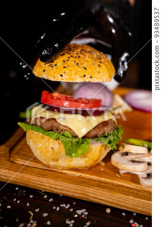 Process of cooking burgers. Cropeed view of chef hands in black gloves with prepared cheeseburger, variety of fillings and ingridients on wooden desk. Catering and Good Quality Fastfood concept 93489537