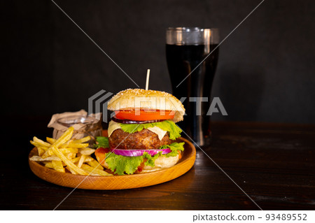 Delicious hamburger with cola and potato fries on a wooden table with a dark brown background behind. Fast food concept. Delicious hamburger with cola and potato fries on a wooden table with a dark brown background behind. Fast food concept. 93489552