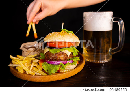 tasty hamburger burger sandwich with french fries and ketchup on a wooden tray a glass of cool beer with foam fast food black background. hand dips french fries in ketchup tasty hamburger burger sandwich with french fries and ketchup on a wooden tray a glass of cool beer with foam fast food black background. hand dips french fries in ketchup 93489553