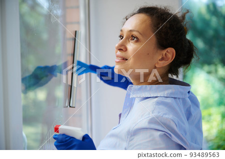 Close-up of multi-ethnic pretty woman, maid, housewife, homemaker spraying detergent and removing stains while washing windows in the veranda. Housekeeping. Household. Chores. Spring cleaning concept Close-up of multi-ethnic pretty woman, maid, housewife, homemaker spraying detergent and removing stains while washing windows in the veranda. Housekeeping. Household. Chores. Spring cleaning concept 93489563