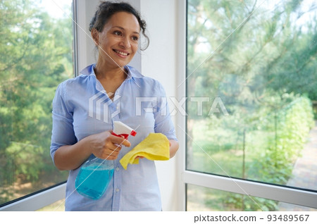 Beautiful multi-ethnic woman housewife, homemaker, maid wearing blue casual shirt, spraying detergent on a yellow cloth while washing panoramic windows in veranda. Spring cleaning and housekeeping 93489567