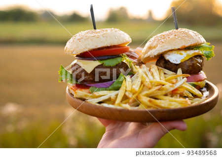 Two hamburgers with beef burger cutlet, fried onion, ketchup sauce and cheese in traditional buns, served on wood chopping board. A hand holds a tray in the open air of the sunset 93489568