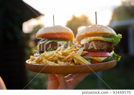 Two hamburgers with beef burger cutlet, fried onion, ketchup sauce and cheese in traditional buns, served on wood chopping board. A hand holds a tray in the open air of the sunset Two hamburgers with beef burger cutlet, fried onion, ketchup sauce and cheese in traditional buns, served on wood chopping board. A hand holds a tray in the open air of the sunset 93489570