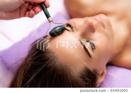 Facial massage. Side view european woman getting massage with jade face roller gouache in spa salon Facial massage. Side view european woman getting massage with jade face roller gouache in spa salon 93491005