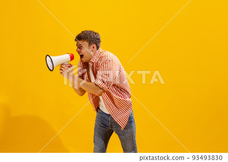 Portrait of young man in checkered shirt posing expressively shouting in megaphone isolated over yellow studio background Portrait of young man in checkered shirt posing expressively shouting in megaphone isolated over yellow studio background 93493830