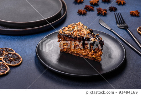 A piece of fresh delicious cake with nuts and chocolate on a black plate against a dark blue background A piece of fresh delicious cake with nuts and chocolate on a black plate against a dark blue background 93494169