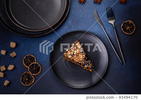 A piece of fresh delicious cake with nuts and chocolate on a black plate against a dark blue background A piece of fresh delicious cake with nuts and chocolate on a black plate against a dark blue background 93494170