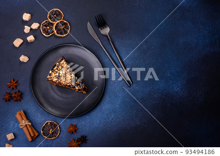 A piece of fresh delicious cake with nuts and chocolate on a black plate against a dark blue background 93494186