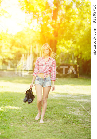 Beautiful young blonde woman with denim shorts and checked shirt walking in the park 93505710