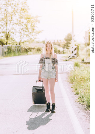 Beautiful young blonde girl smiling on the sunny road with suitcase luggage . Traveling the world with hitchhiking. Beautiful young blonde girl smiling on the sunny road with suitcase luggage . Traveling the world with hitchhiking. 93505711