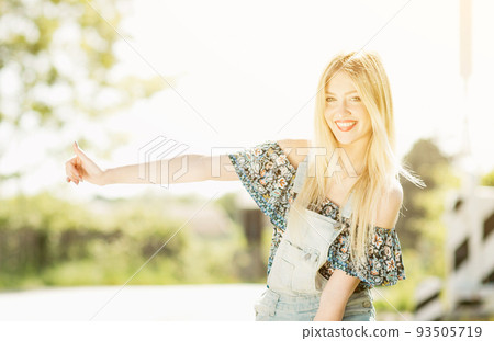 Beautiful young blonde girl smiling on the sunny road with suitcase luggage . Traveling the world with hitchhiking. 93505719