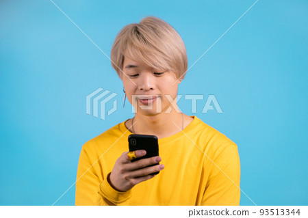 Handsome smiling korean man using smartphone on blue studio wall. Blond guy in yellow outfit using 93513344
