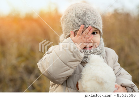 A little two-year-old girl with a soft bear toy covers one eye with her hand and smiles sweetly. Portrait of a child in nature on an autumn day. Problems of children's vision 93515596