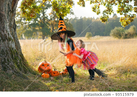 Two two-and five-year-old girls are laughing merrily sitting on a broom under a large oak tree trunk at sunset on Halloween day. Carnival costumes. Kids joyful laughter. halloween sales. background Two two-and five-year-old girls are laughing merrily sitting on a broom under a large oak tree trunk at sunset on Halloween day. Carnival costumes. Kids joyful laughter. halloween sales. background 93515979