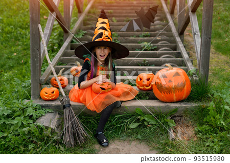 A girl in a witch dress is sitting on the stairs in front of the house with a surprised face on Halloween day with her mouth open. Pumpkins with carved faces stand on old wooden steps. Trick or treat A girl in a witch dress is sitting on the stairs in front of the house with a surprised face on Halloween day with her mouth open. Pumpkins with carved faces stand on old wooden steps. Trick or treat 93515980
