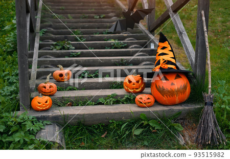 A large pumpkin with a witch's hat next to small pumpkins and a broom stand on the steps of the old staircase in front of the house. Jack o' lantern. Halloween. trick or treating A large pumpkin with a witch's hat next to small pumpkins and a broom stand on the steps of the old staircase in front of the house. Jack o' lantern. Halloween. trick or treating 93515982