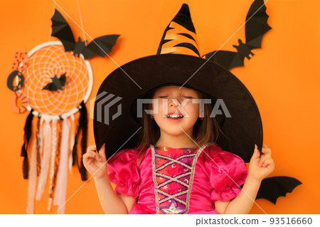 A two-year-old girl in a witch costume on an orange background with bats holds a hat dreamily closing her eyes. Waiting for Halloween Day. Make a wish. trick or treating A two-year-old girl in a witch costume on an orange background with bats holds a hat dreamily closing her eyes. Waiting for Halloween Day. Make a wish. trick or treating 93516660