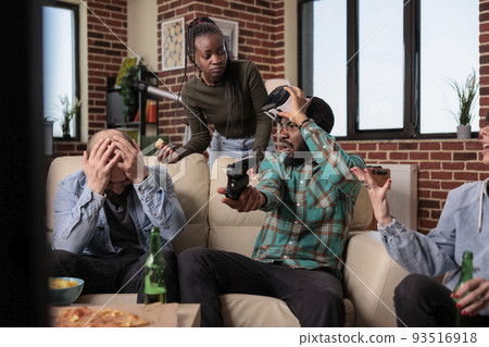 Group of frustrated friends losing video games play with vr glasses and console, drinking bottles of beer. Friends feeling sad about lost gameplay competition, having fun together at gathering. 93516918