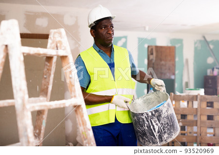 Worker in blue overalls and hardhat dragging various painting tools in room being renovated Worker in blue overalls and hardhat dragging various painting tools in room being renovated 93520969