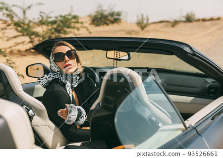 Glamorous blonde in a silk scarf and sunglasses in the driver's seat of a convertible convertible on a hot desert background. Fashion, style, glamour Glamorous blonde in a silk scarf and sunglasses in the driver's seat of a convertible convertible on a hot desert background. Fashion, style, glamour 93526661