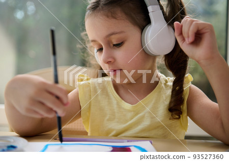 Close-up portrait of Caucasian little girl, adorable preschooler child listening to the music in wireless headphones, holding a paintbrush and painting picture with watercolors in a creative art class Close-up portrait of Caucasian little girl, adorable preschooler child listening to the music in wireless headphones, holding a paintbrush and painting picture with watercolors in a creative art class 93527360