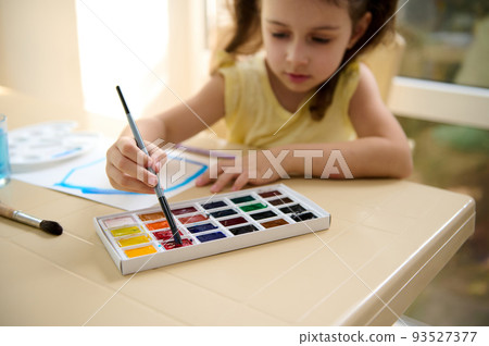 Focus on the hand of a talented little girl, holding paintbrush and mixing colorful paints on a professional watercolor palette. Learning peinture. Art class. Kids education and hobby. Back to school 93527377
