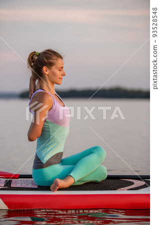 Young woman sitting on paddle board, practicing yoga pose. Doing yoga exercise on sup board, active summer rest Young woman sitting on paddle board, practicing yoga pose. Doing yoga exercise on sup board, active summer rest 93528548