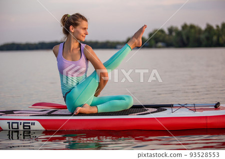 Young woman sitting on paddle board, practicing yoga pose. Doing yoga exercise on sup board, active summer rest Young woman sitting on paddle board, practicing yoga pose. Doing yoga exercise on sup board, active summer rest 93528553