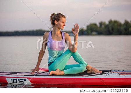 Young woman sitting on paddle board, practicing yoga pose. Doing yoga exercise on sup board, active summer rest Young woman sitting on paddle board, practicing yoga pose. Doing yoga exercise on sup board, active summer rest 93528556