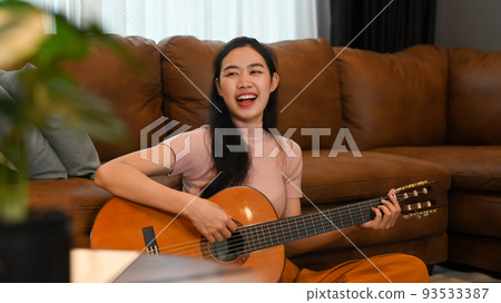 Cheerful young woman playing acoustic guitar, spending leisure time in cozy living room Cheerful young woman playing acoustic guitar, spending leisure time in cozy living room 93533387