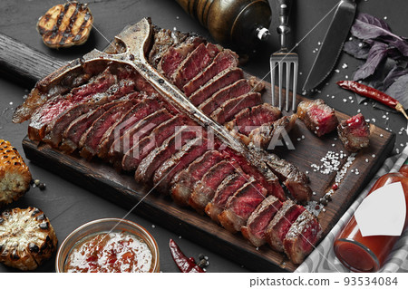 Sliced Steak T-bone lying on wooden board. Grilled vegetables in a pan grill. Black table with gray cloth 93534084