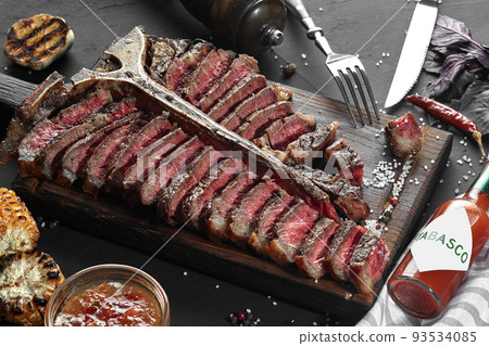 Sliced Steak T-bone lying on wooden board. Grilled vegetables in a pan grill. Black table with gray cloth 93534085