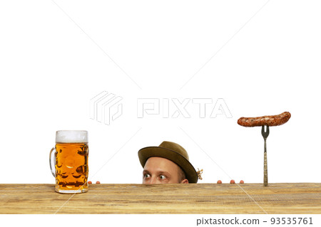 Portrait of young man peeking out table and looking at mug with foamy lager beer and sausage isolated over white background Portrait of young man peeking out table and looking at mug with foamy lager beer and sausage isolated over white background 93535761