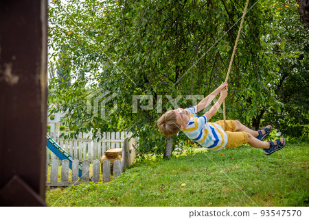 Happy little boy is having fun on a rope swing which he has found while having rest outside city. Active leisure time with children Happy little boy is having fun on a rope swing which he has found while having rest outside city. Active leisure time with children 93547570