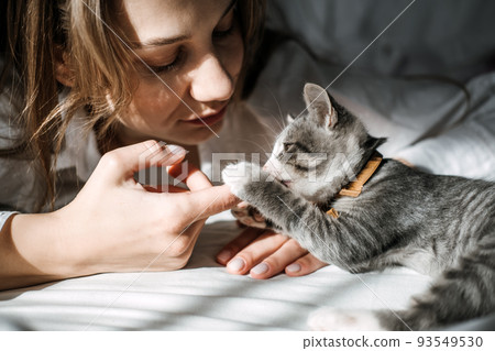 Candid portrait of Young woman is resting with kitten on the bed at home one sunny day. Girl play with outbred kitten. 93549530