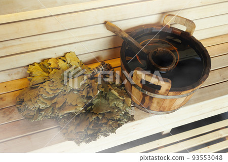 Wooden bucket with water, ladle and a green broom in bath. Sauna Accessories on bench from wooden boards 93553048