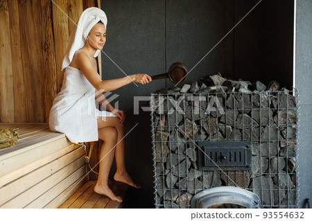 Pretty woman in towel pouring water onto hot stone in sauna 93554632