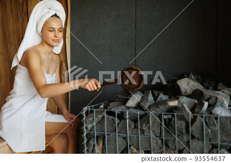 Woman at sauna pours water on a hot stones with a wooden ladle Woman at sauna pours water on a hot stones with a wooden ladle 93554637