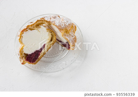 Choux Bun with whipped cream, cherry jam and sugar powder on top, cutted in half on glass plate, white background. Delicate choux pastry dessert. French cream puff. Close-up, copy space 93556339