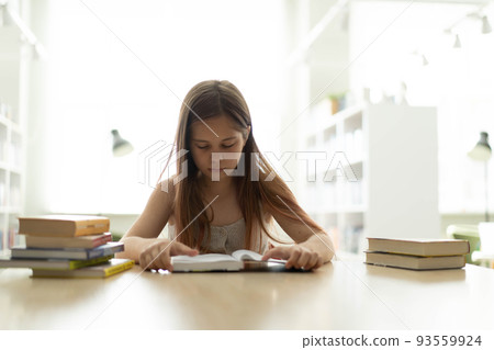 A teenage girl among piles of books. A young caucasian girl is reading a book against the background of shelves. She is surrounded by stacks of books. Book Day. A teenage girl among piles of books. A young caucasian girl is reading a book against the background of shelves. She is surrounded by stacks of books. Book Day. 93559924