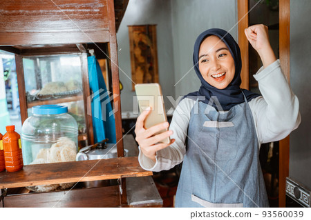 veiled woman wearing an apron happily looking at her cellphone 93560039