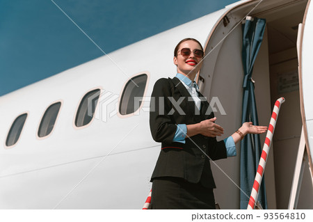 Woman flight attendant standing on airplane stairs at airport and invites passengers on board Woman flight attendant standing on airplane stairs at airport and invites passengers on board 93564810