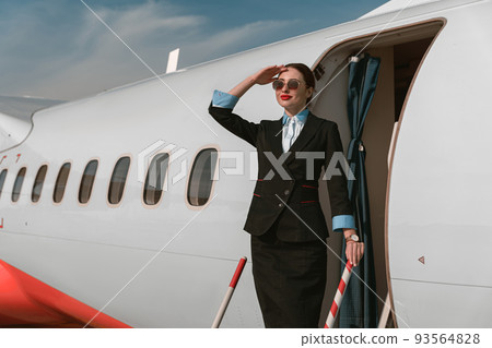 Woman flight attendant in sunglasses standing on airplane stairs at airport and looking away 93564828