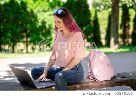 Young girl is studying in the spring park, sitting on the wooden bench and browsing on her laptop Young girl is studying in the spring park, sitting on the wooden bench and browsing on her laptop 93578196
