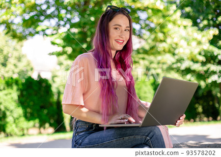 Young girl is studying in the spring park, sitting on the wooden bench and browsing on her laptop 93578202