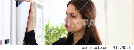 Beautiful woman clerk is looking for correct document on shelf Beautiful woman clerk is looking for correct document on shelf 93579096